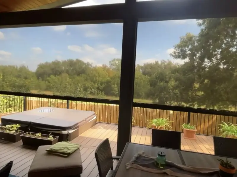 The view from inside a covered deck through lowered mesh toward trees, a hot tub and potted plants.