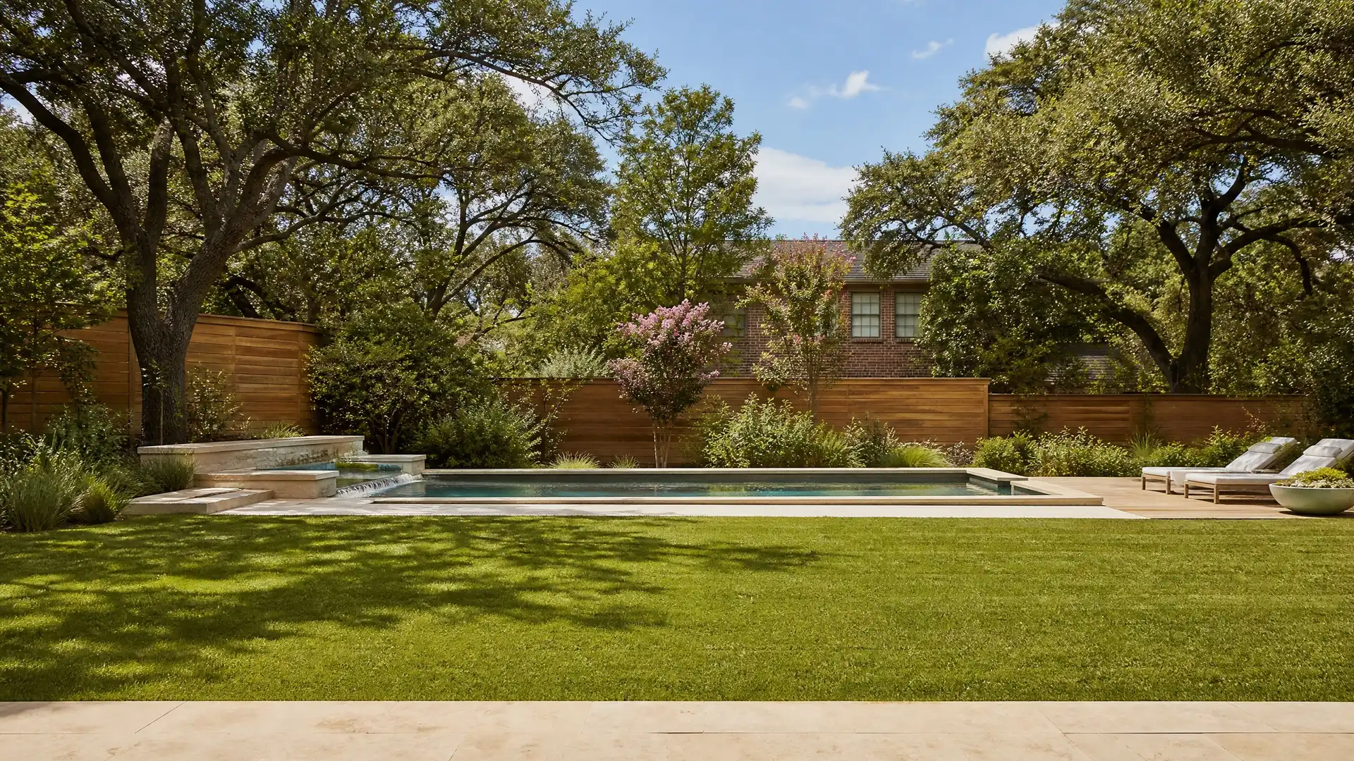 A backyard seen from the patio: pool, live oaks, a crepe myrtle in bloom, a cedar fence, and the neighbor’s two-story brick house with two windows facing the yard.