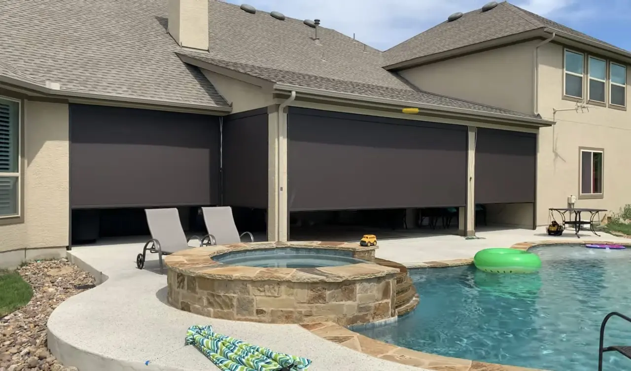A two-story stucco and stone home with four dark motorized screens partly lowered over a patio, a pool and stone spa in front.