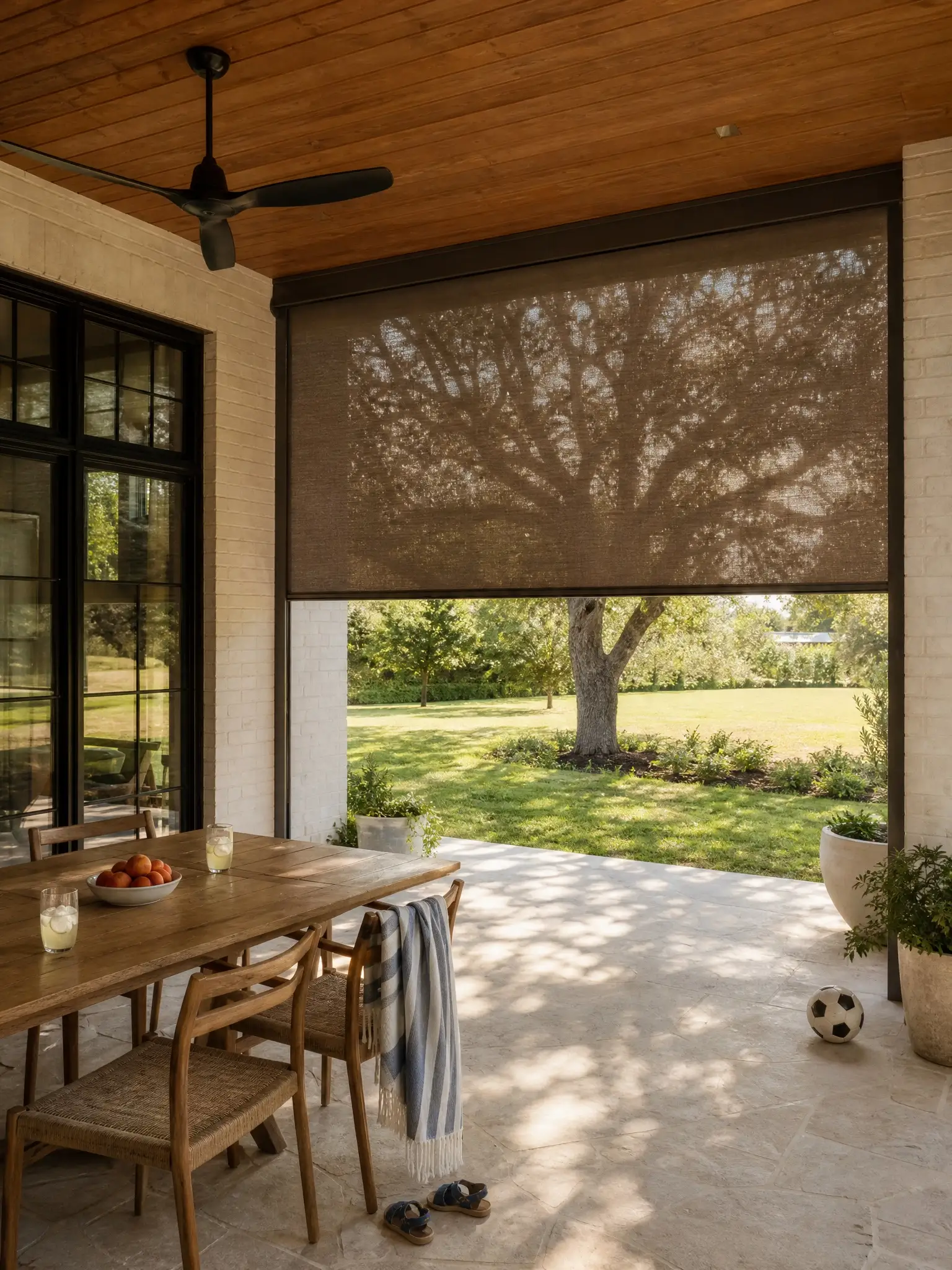 A white-brick farmhouse patio with a bronze shade lowered halfway, lemonade and peaches on the table, a soccer ball on the floor.